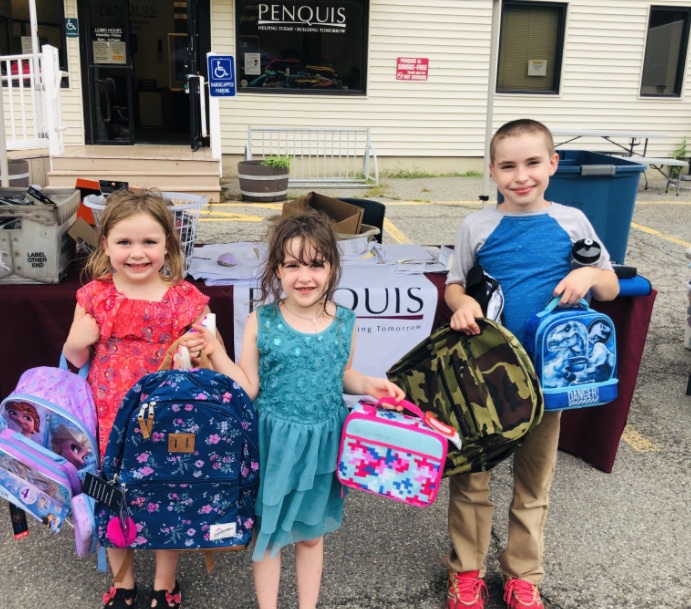 Three young girls smiling and holding colorful backpacks, including Barbie and tie-dye designs, after receiving school supplies from the We've Got Your Back (WGYB) program.