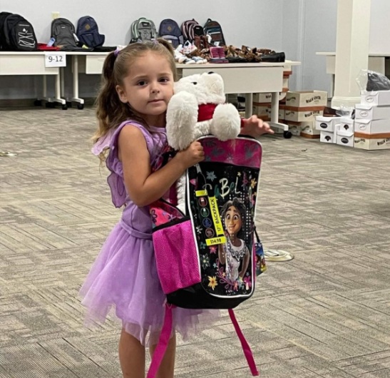 A young girl in a purple dress holds a character-themed backpack and a white stuffed animal, surrounded by tables of backpacks and shoes at a WGYB school supply distribution event.